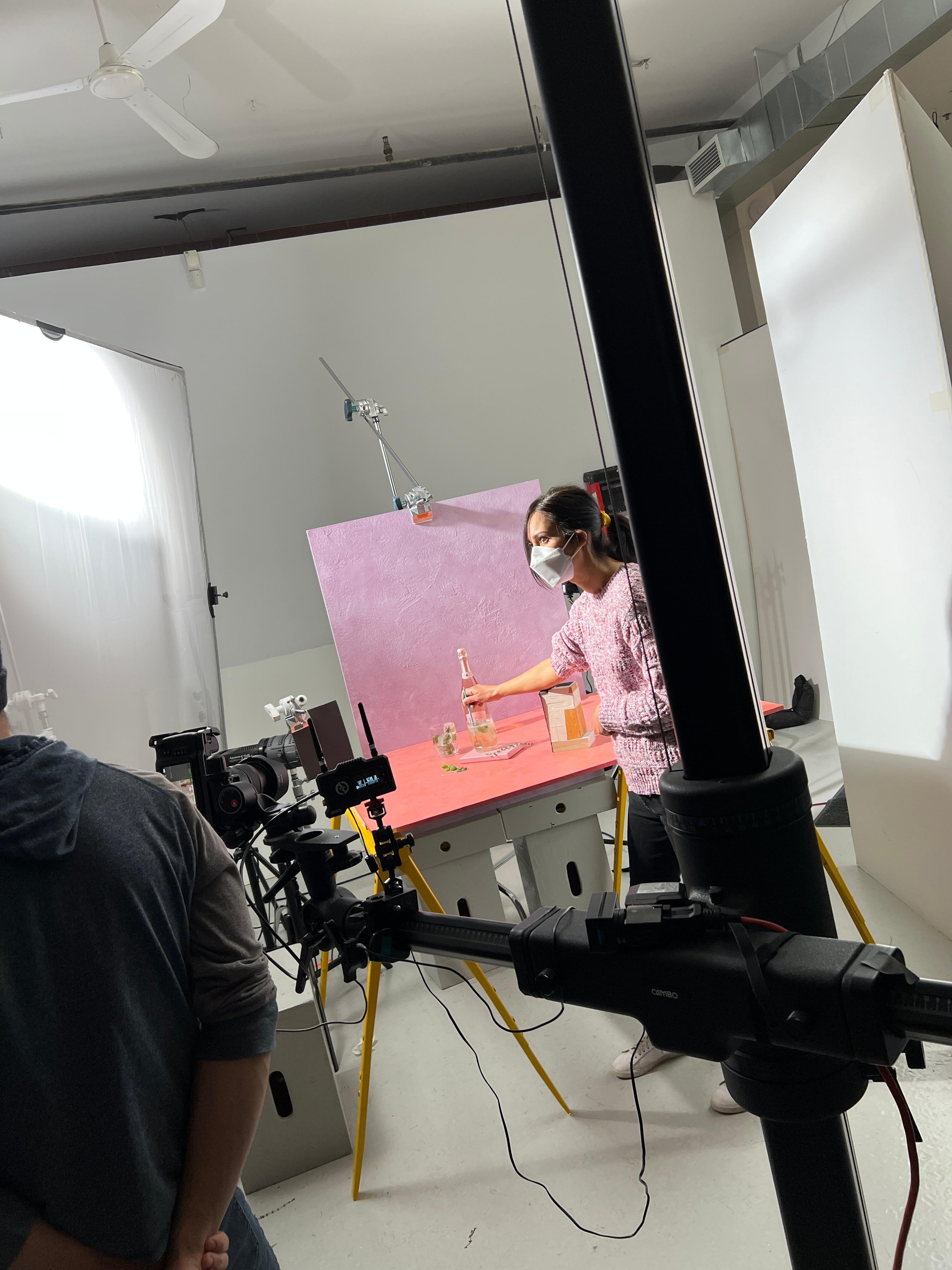Young woman in a pink sweater on a photo studio set. The set has a pink background and she is styling some wine and glasses. There is a lot of studio camera gear around the set.