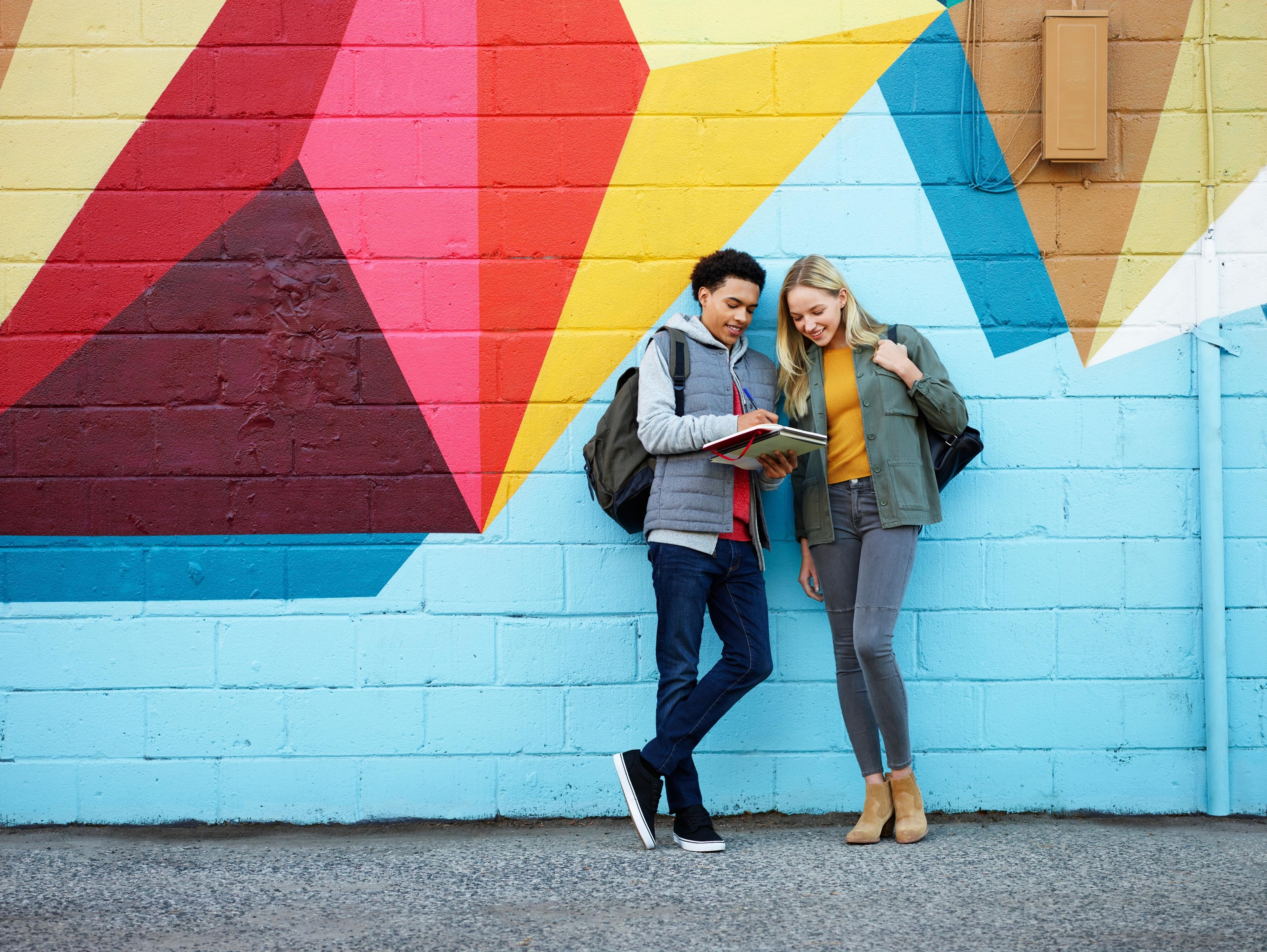 Two smiling teens leaning against a brick wall with a colourful mural looking at a notebook